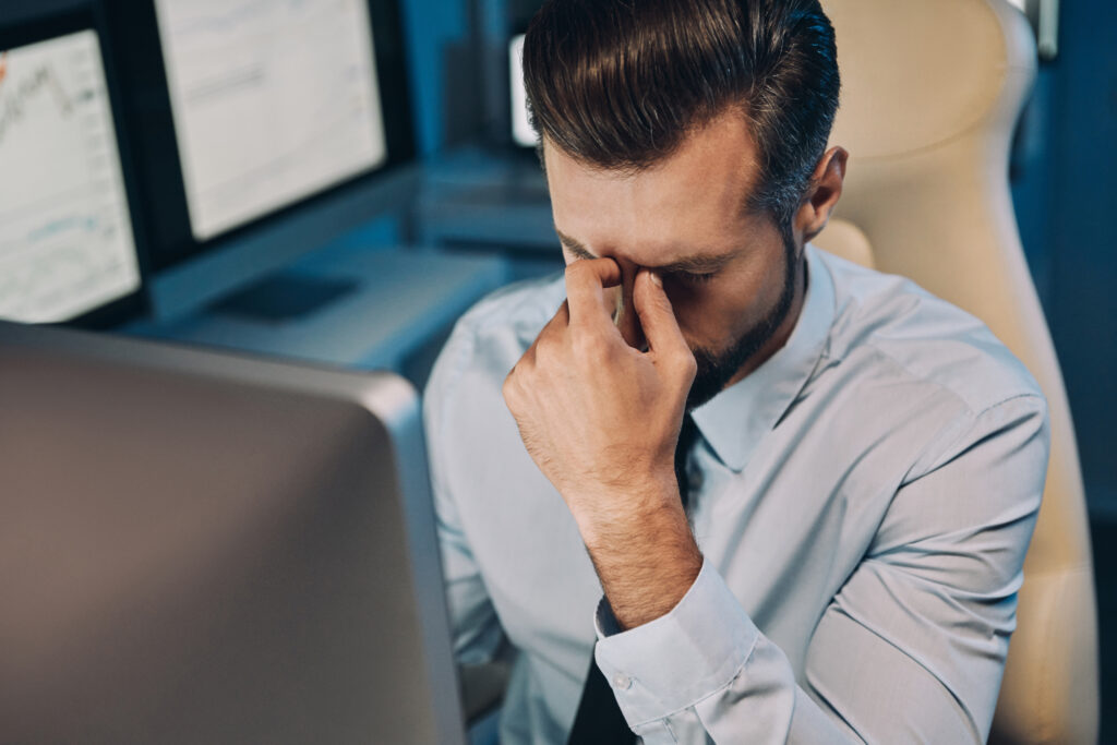 Homem de camisa social azul e gravata preta está sentado em frente a um computador com a cabeça abaixada e a mão apoiada no nariz, com uma expressão de estresse e cansaço pelos erros cometidos na hora de abrir empresas e negócios.