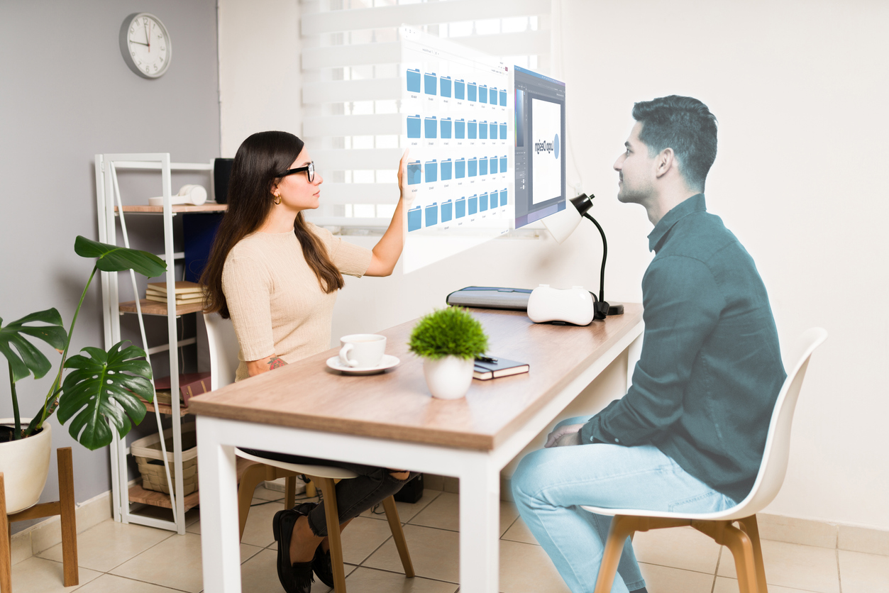 Uma mulher de óculos e blusa bege está sentada em uma mesa e interagindo com um holograma de um homem. A mulher apresenta alguns aquivos da empresa por meio da tecnologia do metaverso