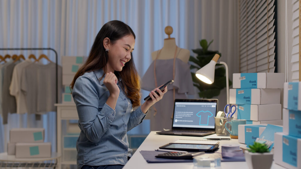 Uma mulher de camisa azul está em uma sala com manequim e roupas, ela está segurando um celular e olhando para a tela enquanto sorri, na mesa tem um computador e algumas ciaxas para enviar produtos