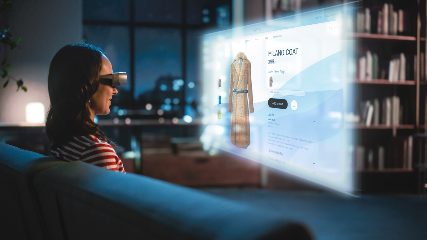 Uma mulher está sentada em um sofá usando óculos de tecnologia de realidade virtual, na frente da mulher aparece uma imagem de uma tela com um casaco para ser comprado em um site