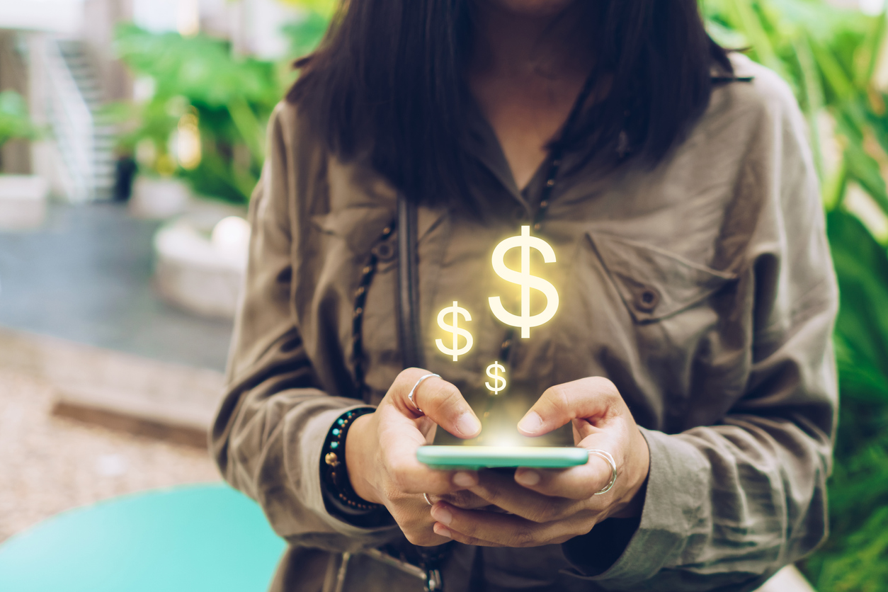 Uma mulher aparece carregando um celular e saem da tela do aparelho símbolos de dinheiro e cifrão