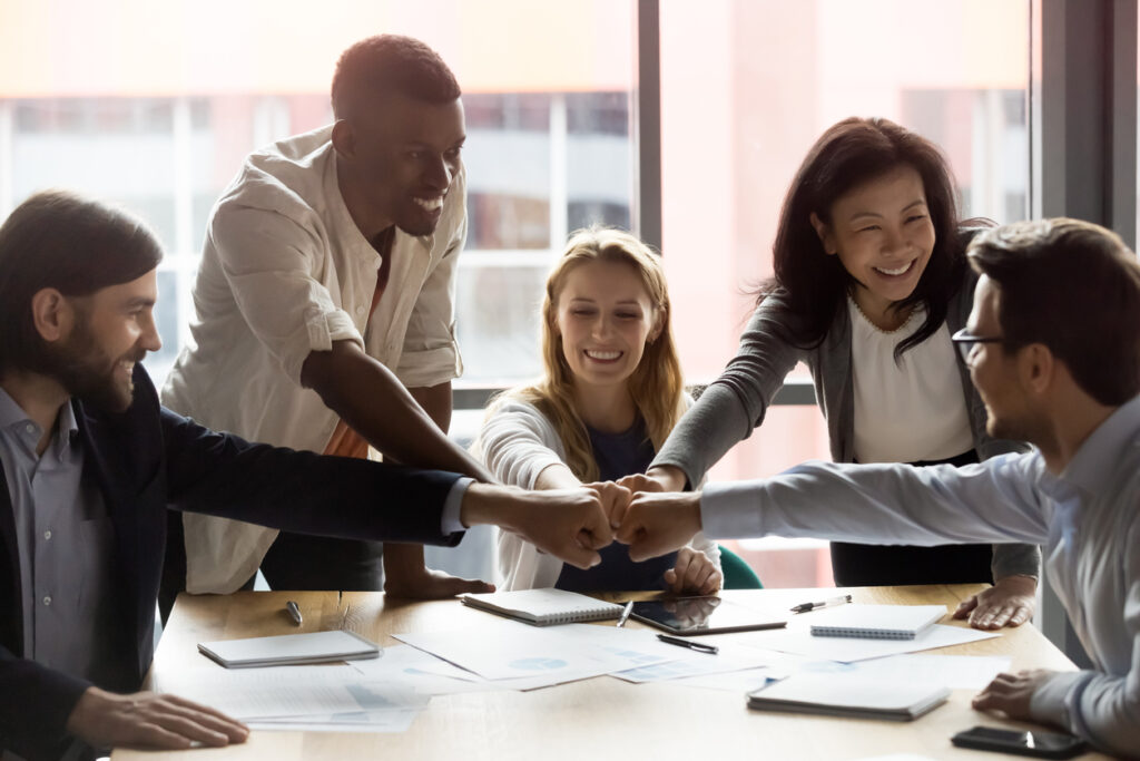 Um grupo de pessoas est reunido em uma mesa durante o trabalho, todos esto sorrindo e eles juntam as mos no centro em sinal de trabalho em equipe. Importncia do trabalho de endomarketing nas empresas.