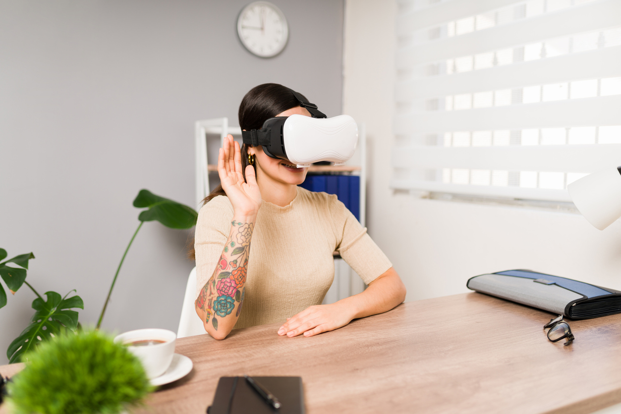 Uma mulher de blusa bege est usando culos de realidade virtual, ela est sentada em uma mesa e o brao direito levantado enquanto ela interage virtualmente. Metaverso e o seu impacto para os negcios na era atual.