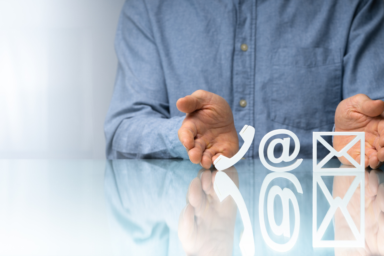 Um home de camisa azul está com as duas mãos em cima de uma mesa delimitando um espaço, entre as mãos estão ícones de um telefone, de um @ de e-mail e de um envelope que simboliza mensagem de telefone
